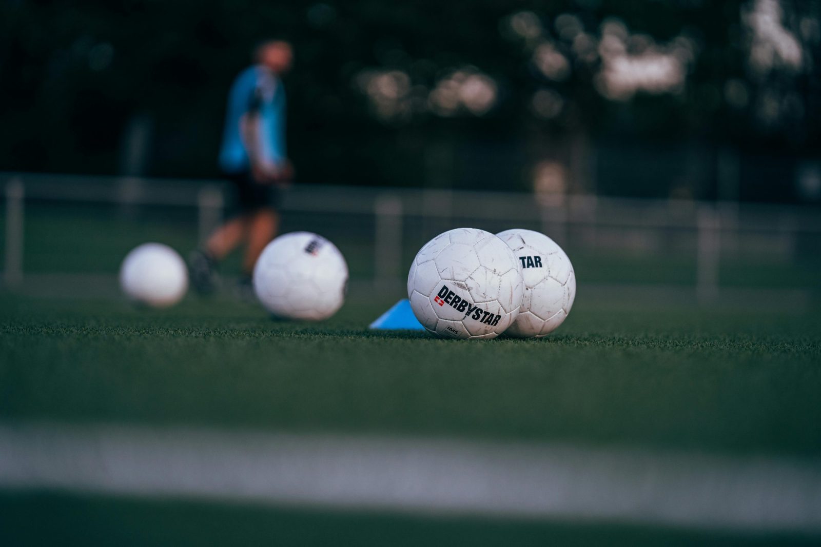 Soccer balls on a green field during a training session, blurred coach in background.